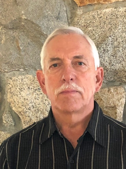 Portrait of an individual with short gray hair, standing against a stone wall, wearing a collared black and gray striped shirt. The person maintains a neutral expression.