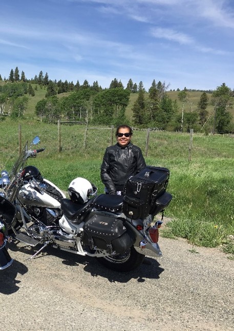 Person in black leather jacket standing beside a Harley-Davidson motorcycle in a scenic outdoor setting with green hills and sparse trees.