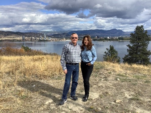 Two people standing together in an open field with a scenic backdrop of a large lake and mountains under a cloudy sky.