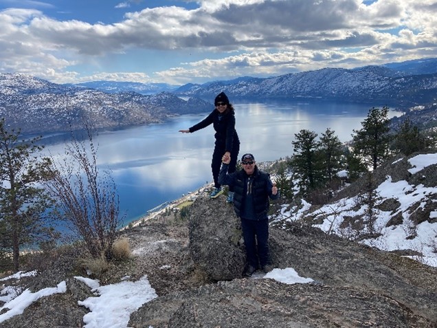 Two people standing on a rocky overlook with a panoramic view of a snow-dusted landscape and a large lake surrounded by mountains. One person is balancing on one foot on a rock, and the other is standing beside the rock with one hand raised.