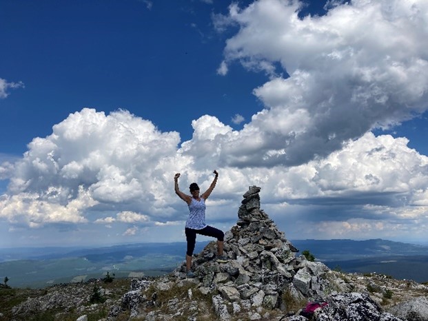 A person is joyfully raising their arms on top of a rock pile at a mountain summit, under a vast blue sky dotted with fluffy white clouds. The landscape below includes distant hills and a clear horizon.