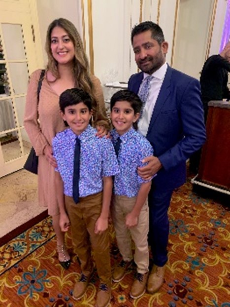 A woman with long hair in a formal pink dress and a man with dark hair and beard in a blue suit stand behind two young boys in blue short-sleeve dress shirts, ties and khaki pants