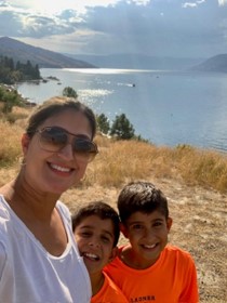 A woman with sunglasses and her hair tied back wearing a white shirt takes a selfie with twin boys both wearing orange shirts in front of a lake with dry grasses in the background