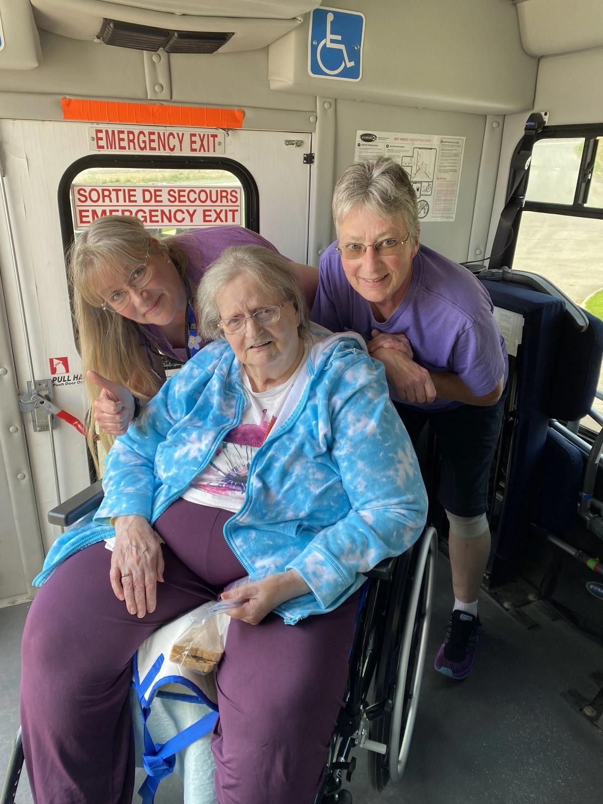 Three individuals, one seated in a wheelchair, smiling inside a bus near an emergency exit sign.