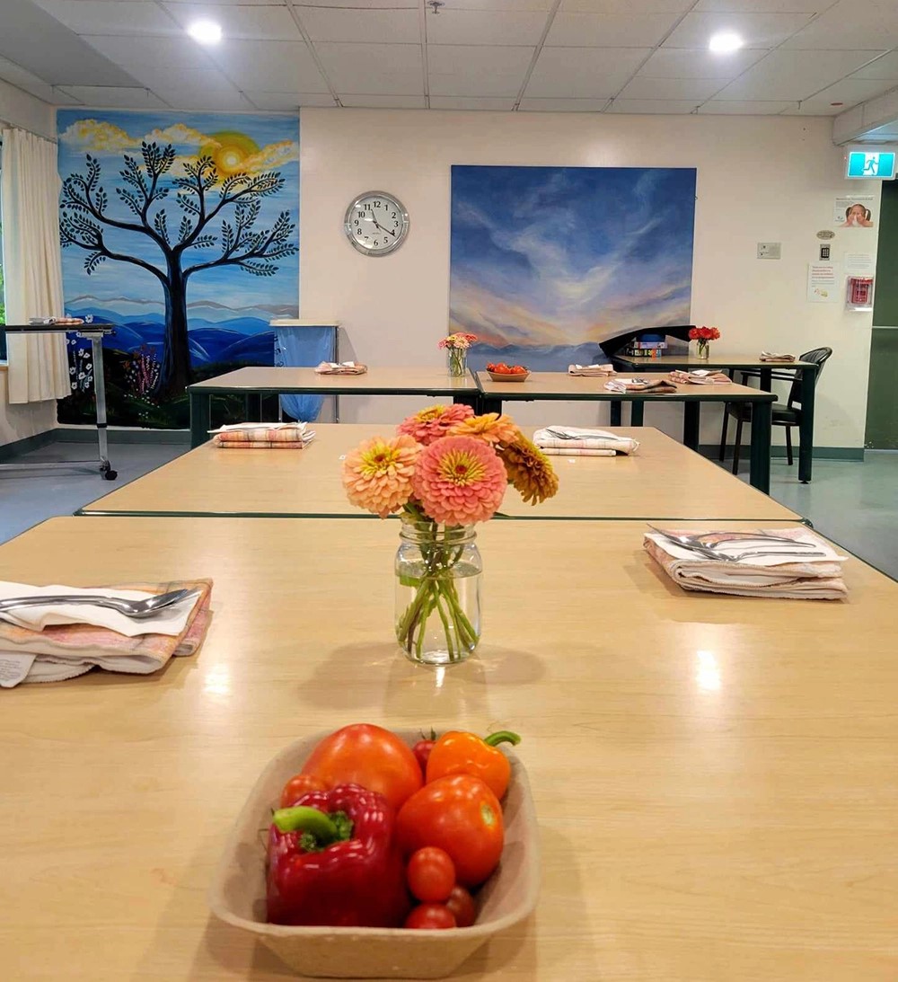 A well-lit dining area with neatly arranged tables, each set with placemats and utensils. A vibrant bouquet of flowers in a vase and a basket of colorful bell peppers are in the foreground. Artistic representations of a tree and a vivid sky painting decorate the walls. A wall clock shows the time.