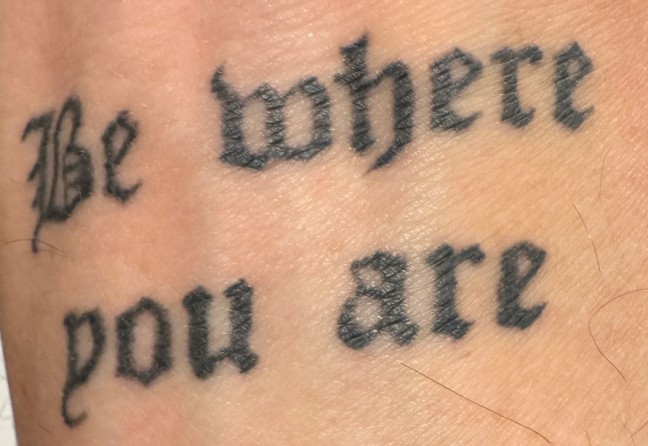 A close-up image of a tattoo on skin reading "Be where you are" in cursive font.