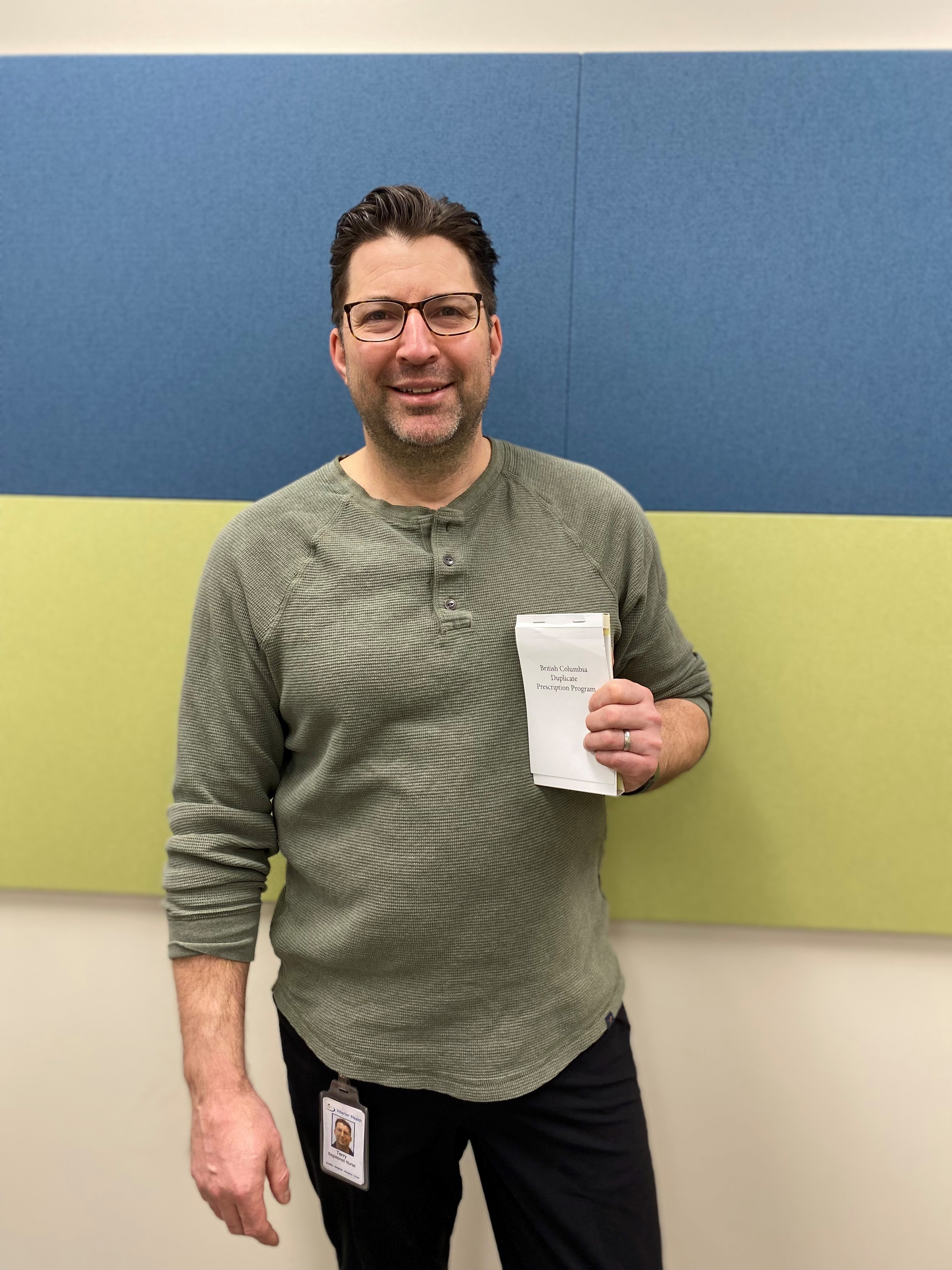 A smiling person wearing glasses standing in front of a blue, green and white striped wall holds a prescription pad