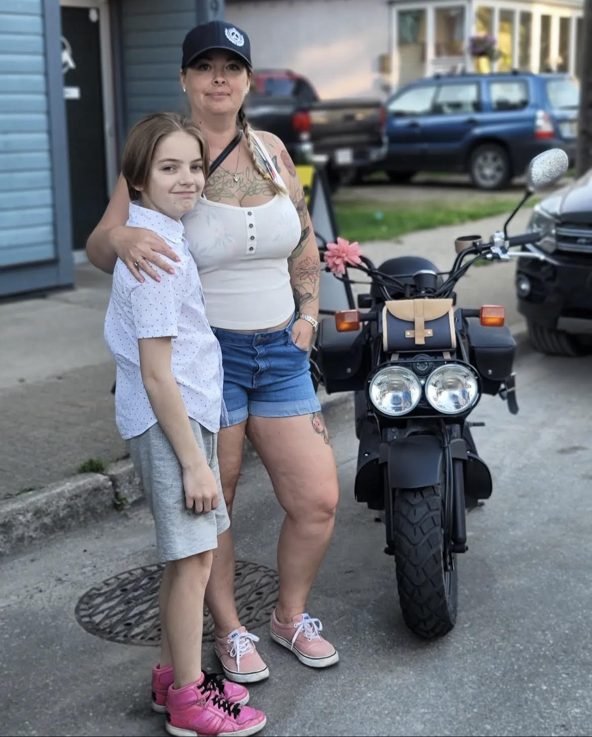 Woman wearing a ballcap, yellow top and jean shorts, one hand in pocket, arm around teenage boy wearing white shirt, shorts and pink running shoes, pose standing next to scooter