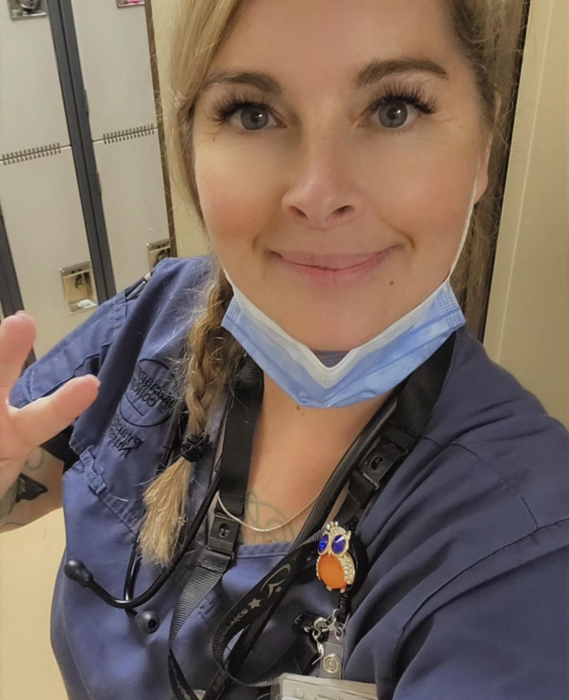 Selfie of blonde woman wearing blue scrubs and blue face mask below chin