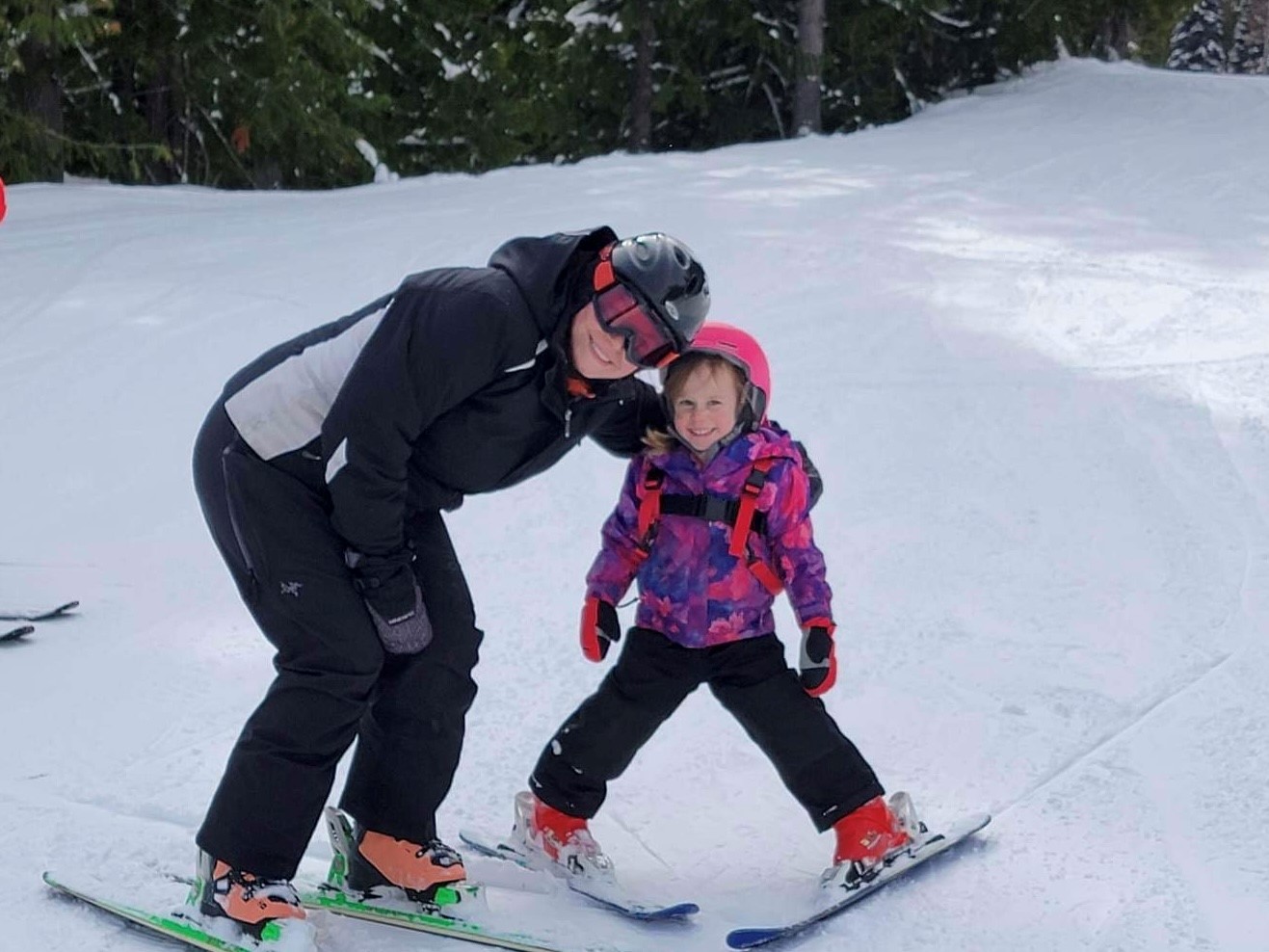 Woman wearing ski goggles, jacket, snow pants, boots and skis with girl wearing pink helmet mittens, standing on snow