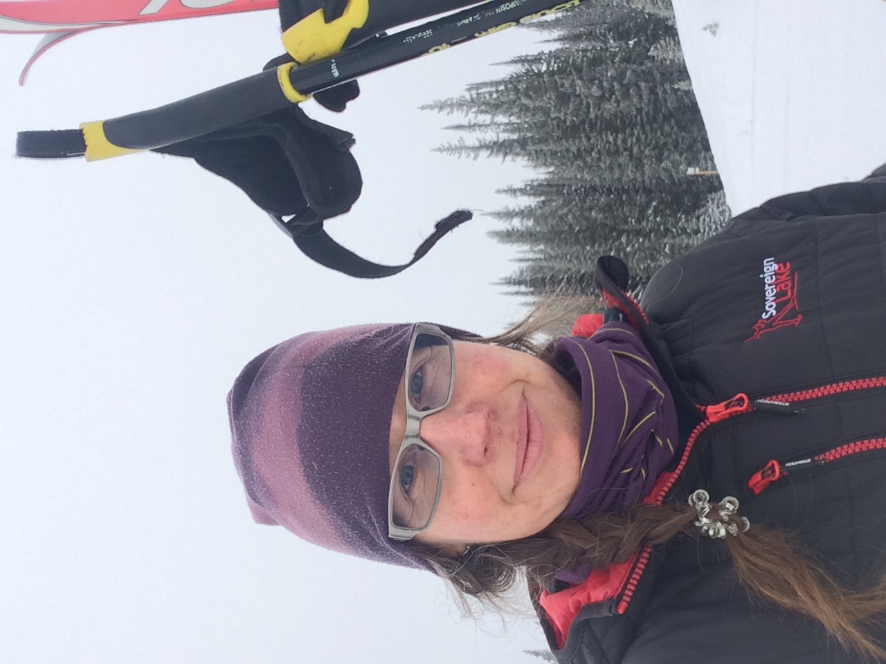 Woman with long hair in a braid, wearing hat, glasses and jacket, holding skis and poles with snow and evergreen forest in background