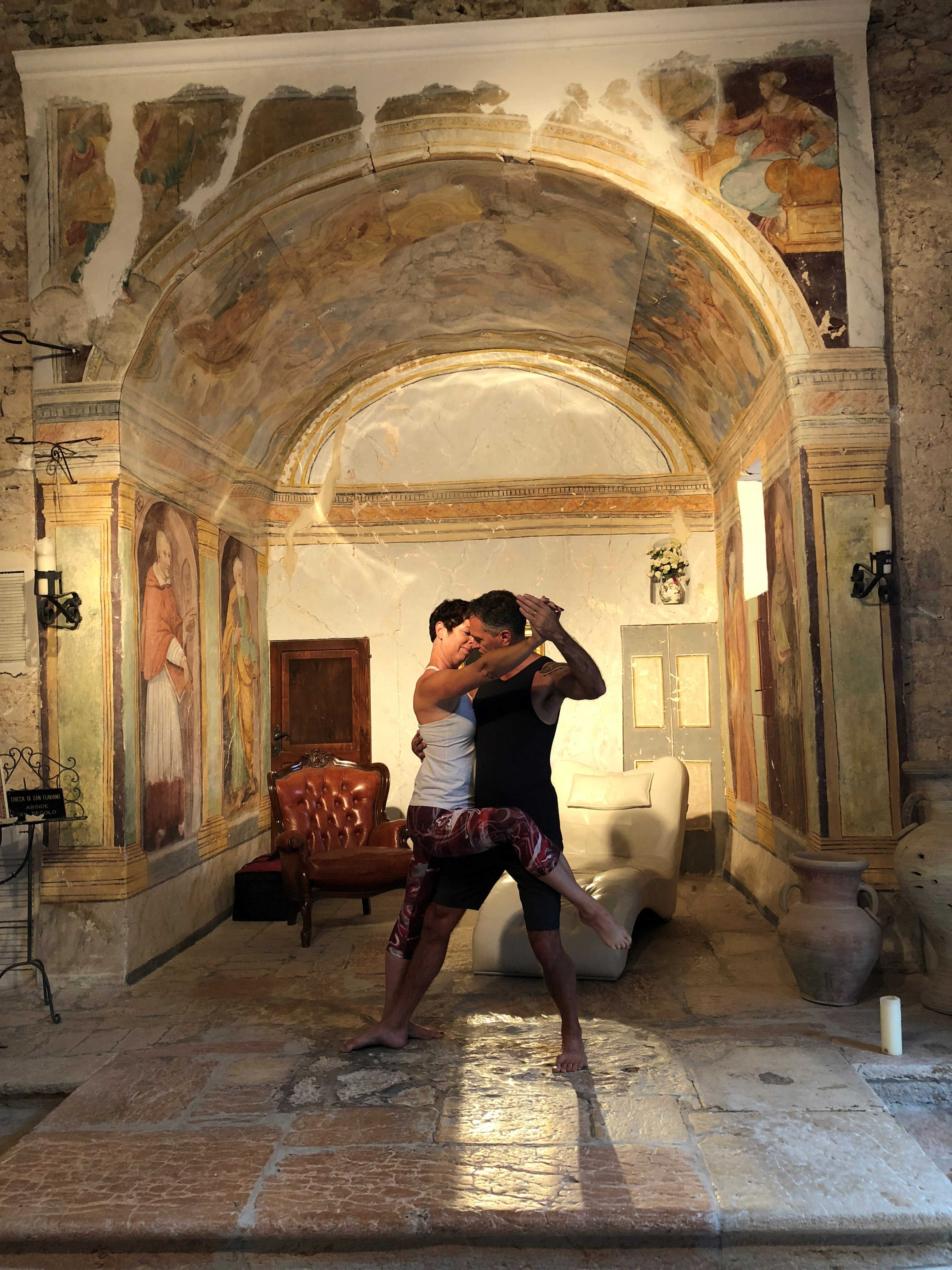 A woman and man in a tango position in front of an ornate archway, with plush chairs (one red, one white) behind.