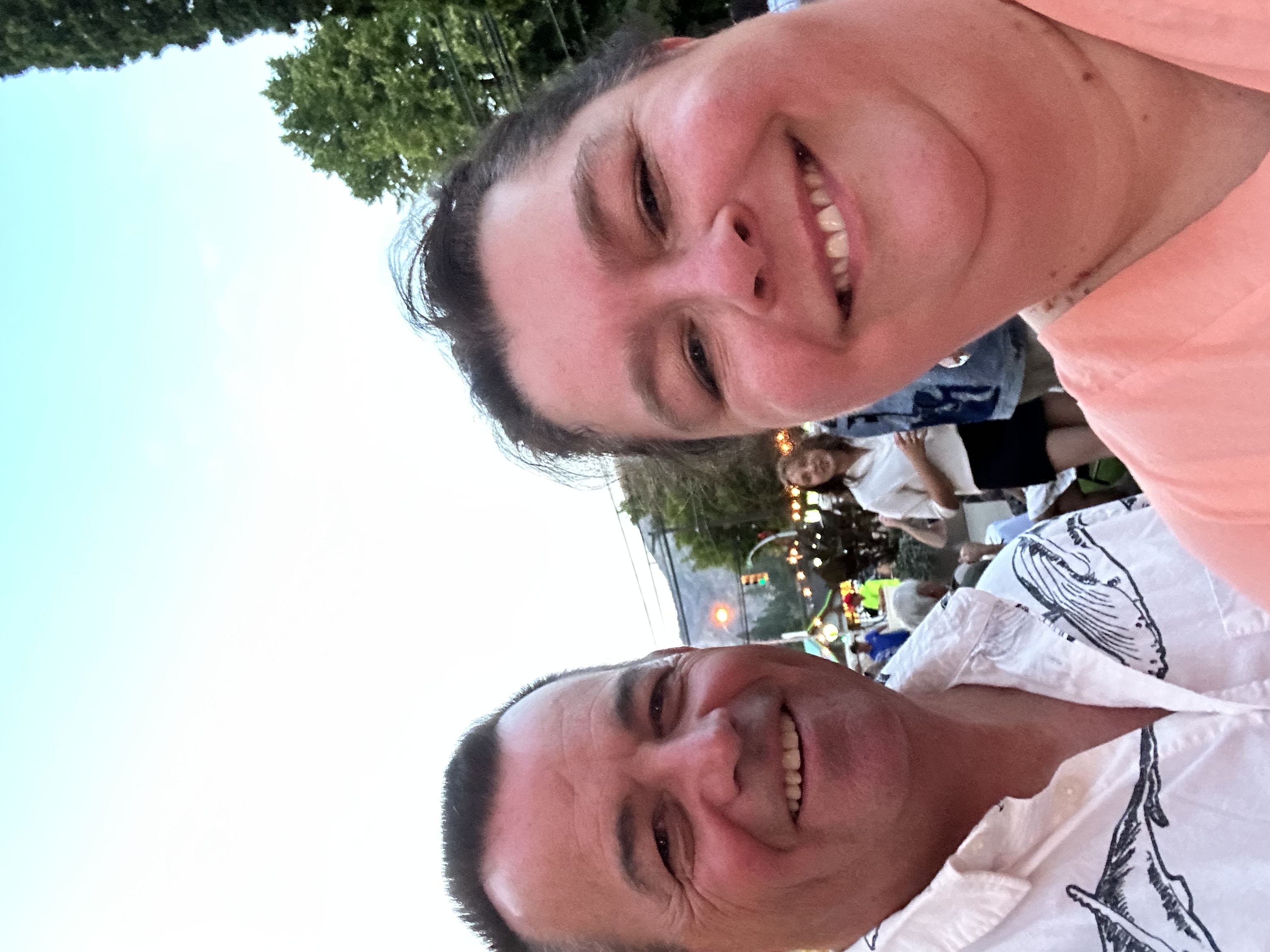 A man and woman take a selfie together outside with a group of people in the background. 