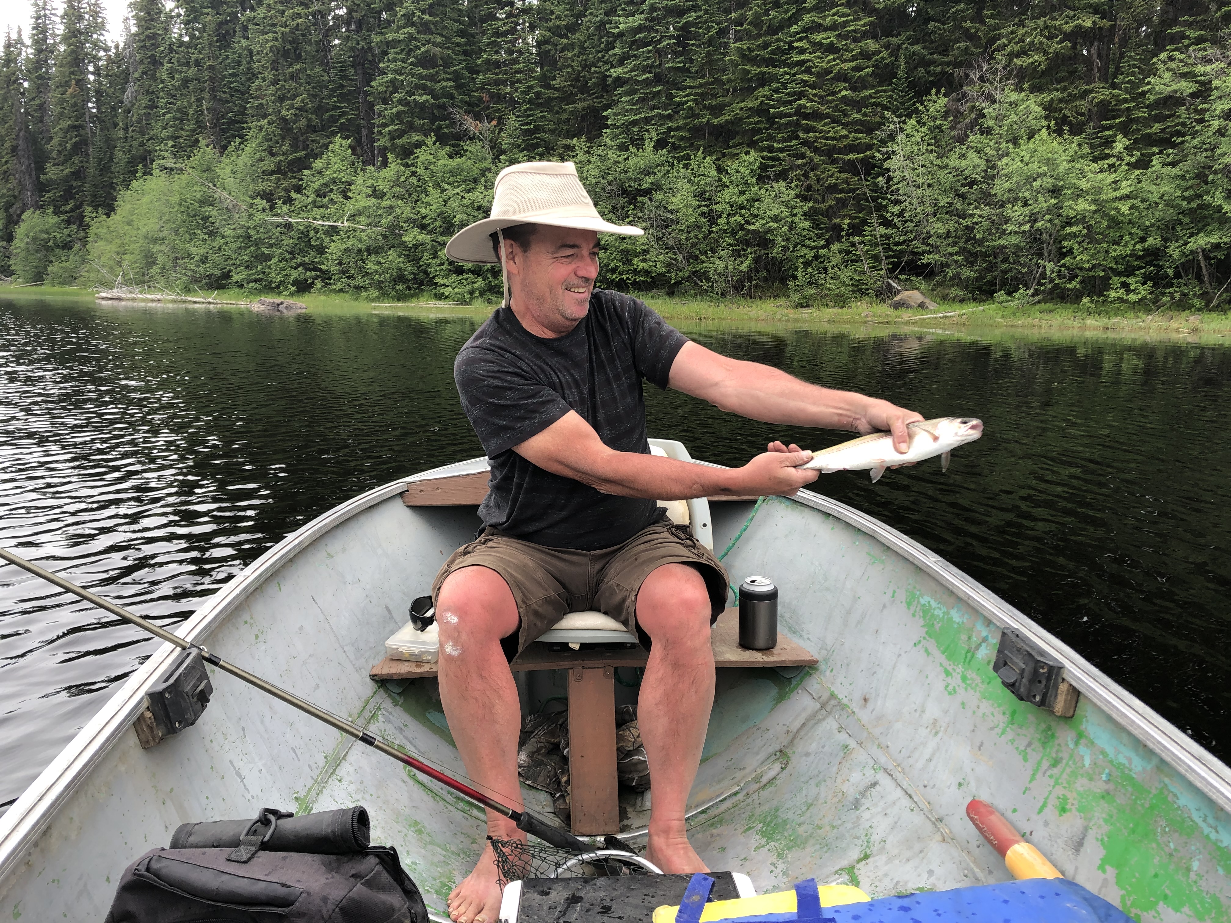 A smiling man sitting in a boat on the water smiles as he holds a fish. There is a fishing rod and fishing gear in the boat.