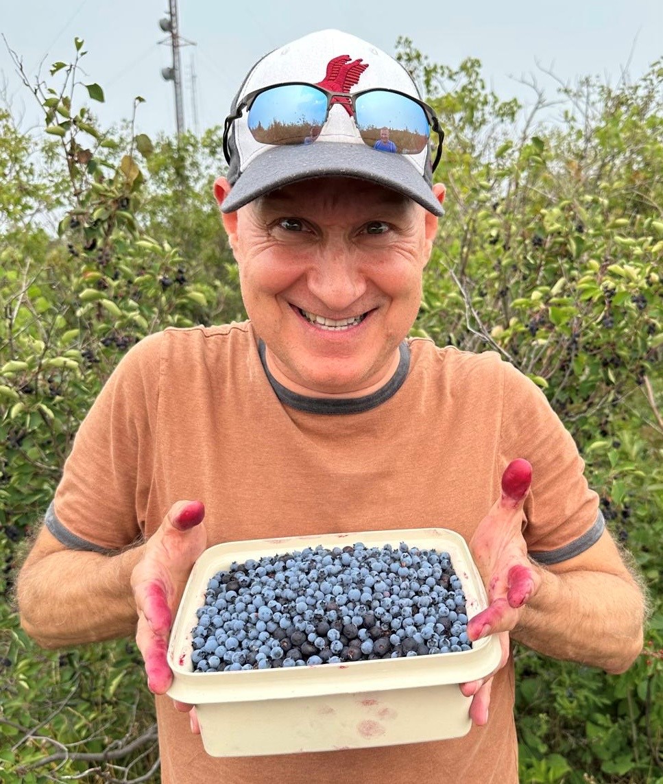 A man standing outside near a bush smiles while holding up a container of blueberries with fingers that have been stained from the fruit.