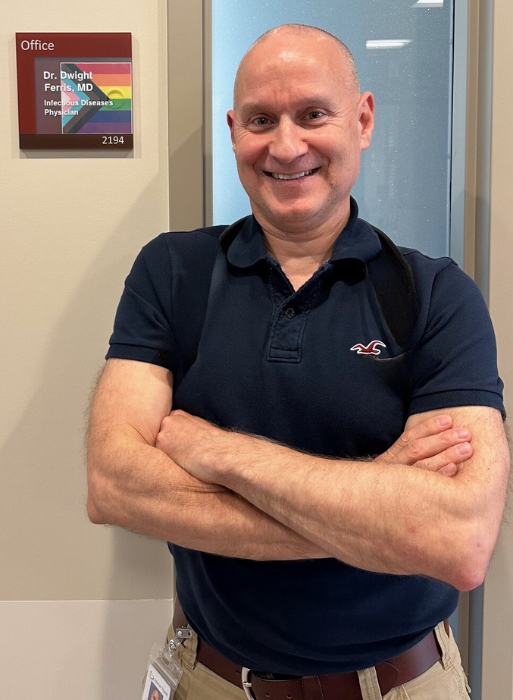 A man wearing a business casual shirt with his arms crossed poses for a photo by an office door with a plaque beside it that features a rainbow flag and text reading Dr. Dwight Ferris, MD, Infectious Diseases Physician.