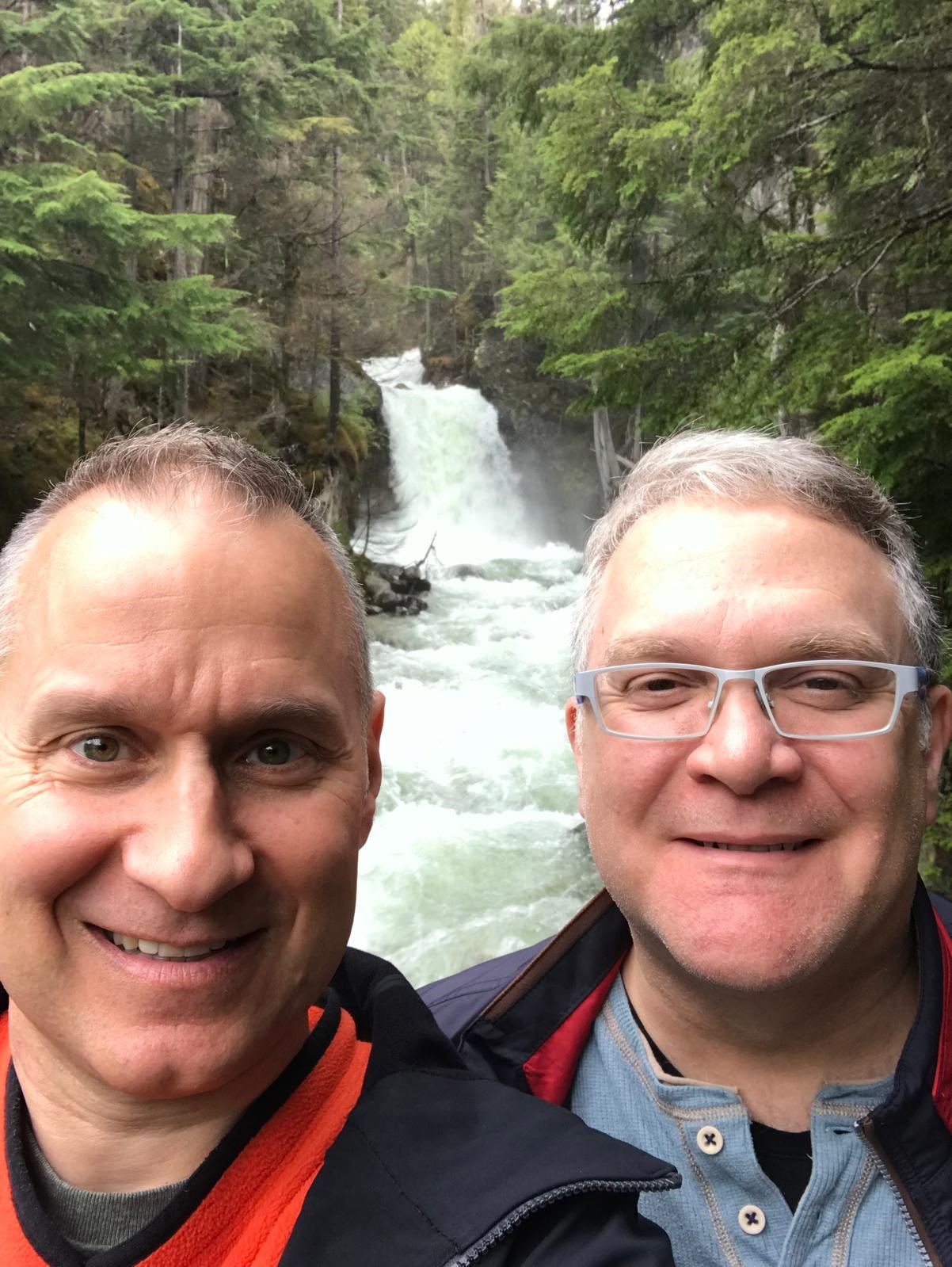 Two men take a selfie with a small waterfall and trees in the background.