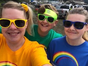 Woman takes a selfie with her son and daughter. She is smiling, has blonde hair tied into a ponytail with a yellow scrunchy and is wearing yellow sunglasses and a yellow shirt with a rainbow on it. Beside her, her son is wearing a green shirt with a rainbow on it with green sunglasses and a green headband. To the right of him, the daughter is wearing a blue rainbow shirt and blue sunglasses.