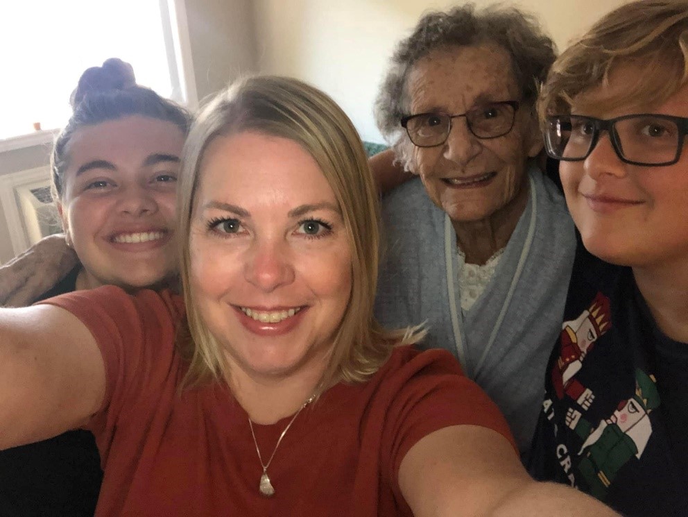 A woman with blonde hair taking a selfie. She is smiling, wearing a red T-shirt. Behind her and to the left in the photo her daughter with brown hair tied in a top knot is smiling. Beside her daughter is her grandmother who is wearing a blue sweater and has short, curly grey hair and black framed glasses. To the right of her grandmother is her son who has short dirty blonde hair and black rimmed glasses and is wearing a black T-shirt.