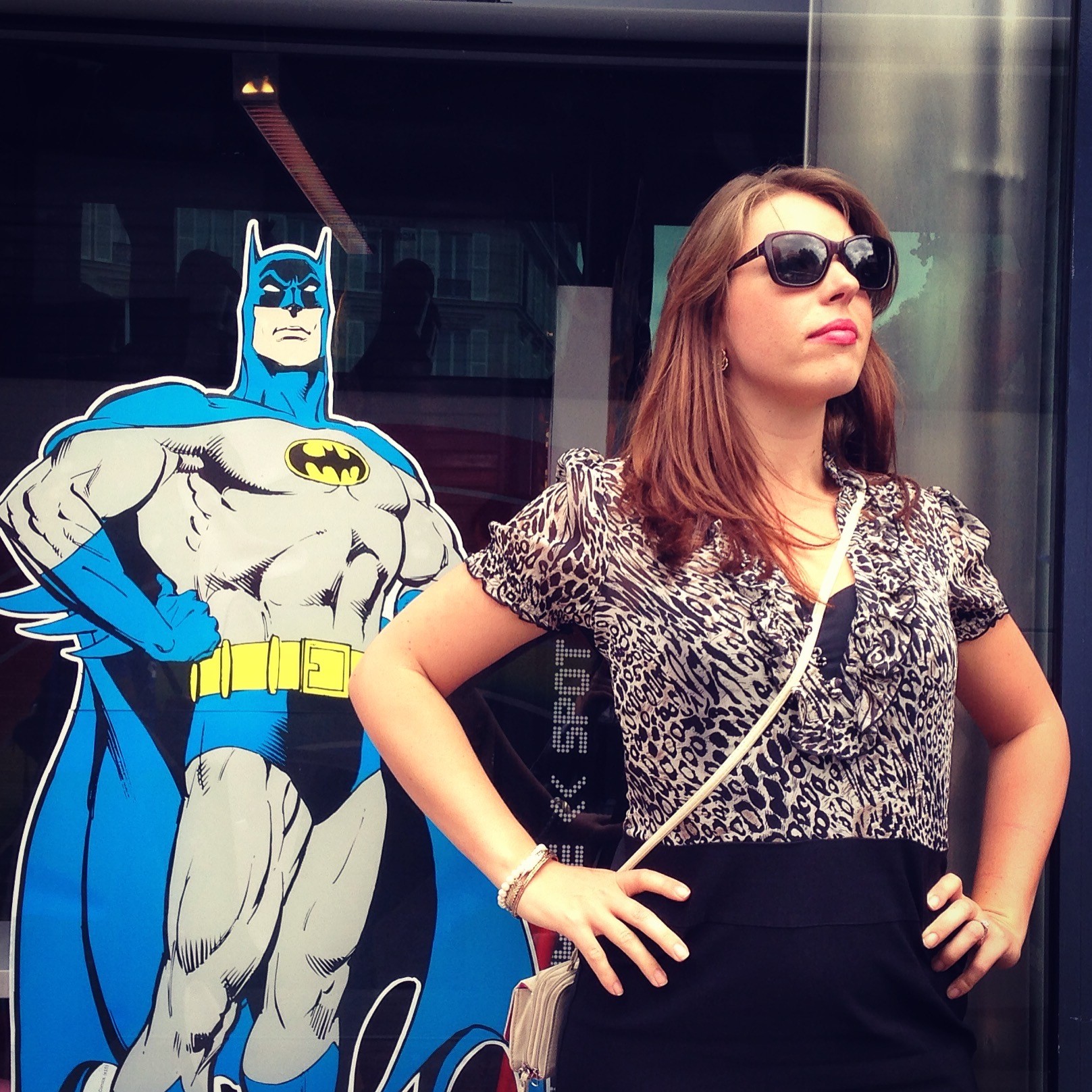 Woman with brown hair wearing sunglasses, short-sleeved black and white blouse, black skirt and beige shoulder bag, poses with hands on hips in front of cardboard cut-out of superhero Batman