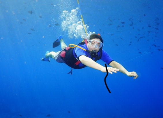 A man in a blue shirt wearing scuba gear blows out bubbles in an underwater picture.