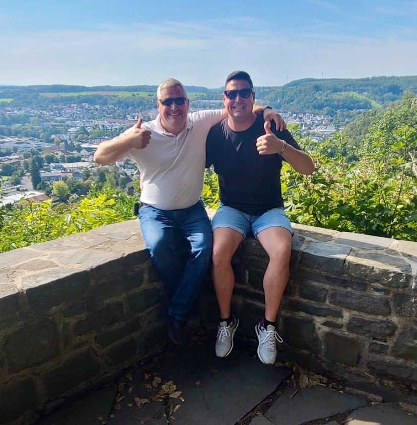 Two men one wearing a white t-shirt and one wearing a blue t-shirt with their arms around another giving the thumbs up sign sit on a stone wall with a lush green city and blue sky in the background.