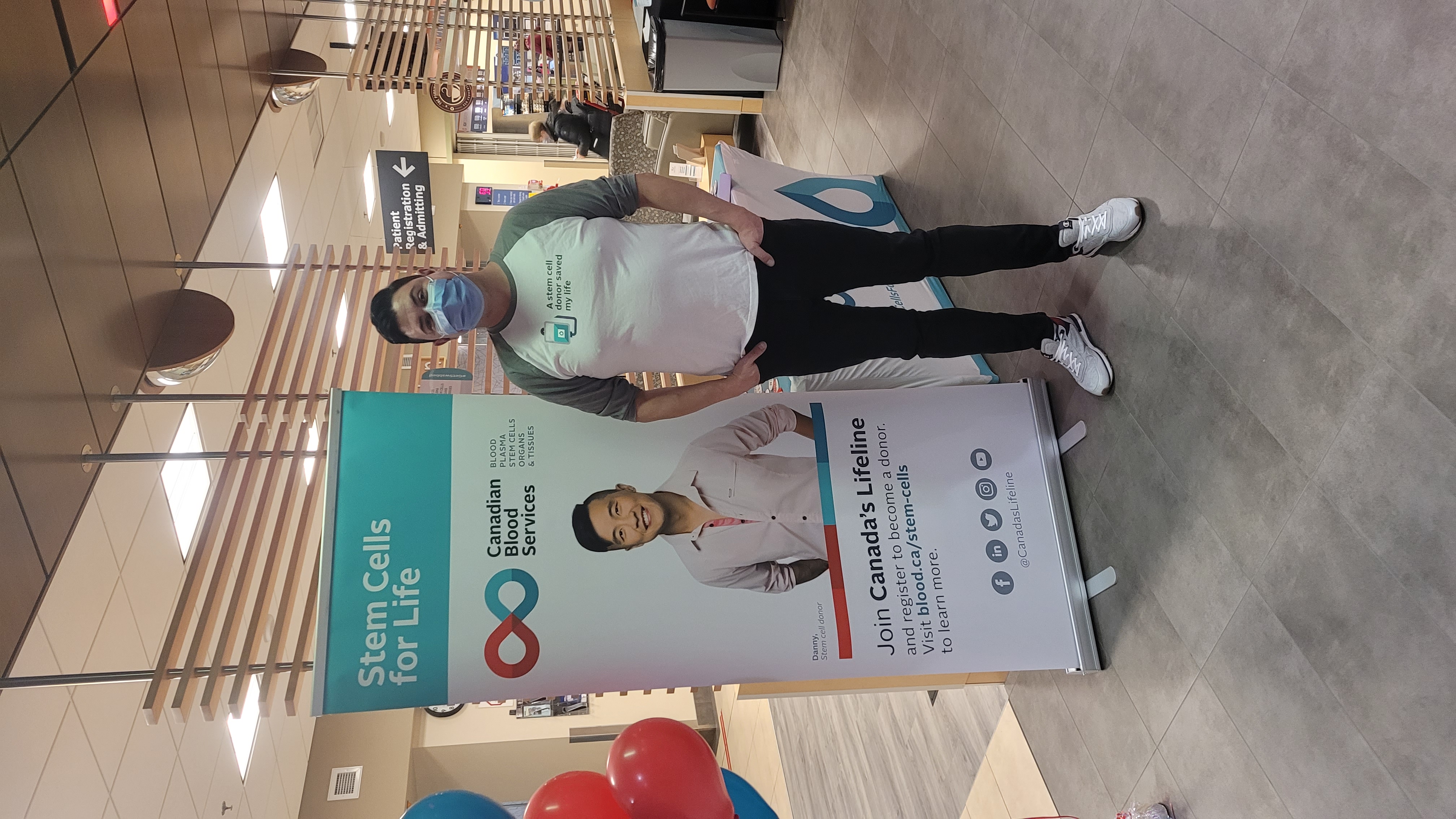 A man standing in a hospital lobby with his hands in his pockets wearing a face mask, white t-shirt with grey sleeves that says “A stem cell donor saved my life” next to a sign that says “Stem Cells for Life” with an image of a smiling man in a white dress shirt.