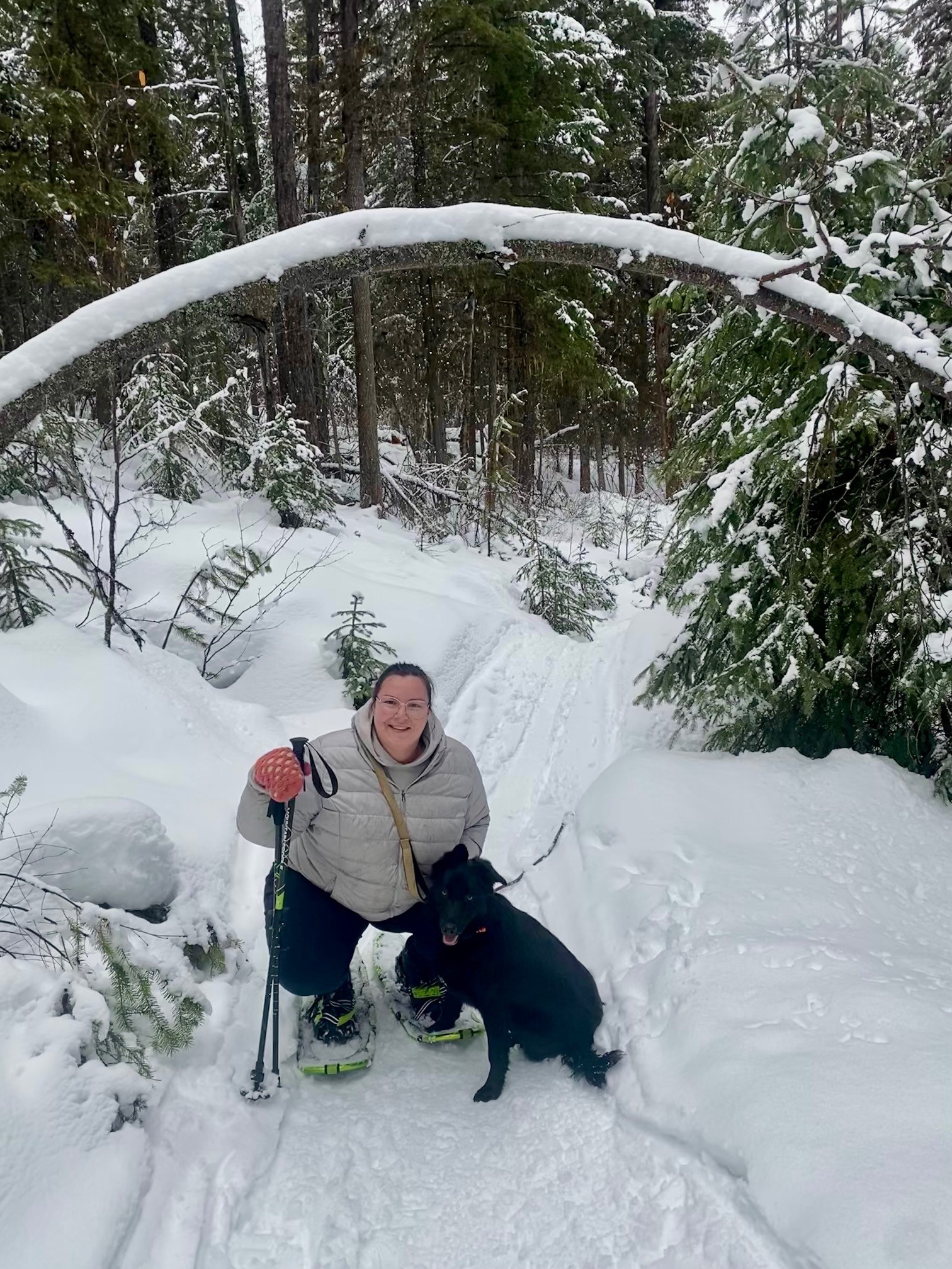 : Woman in light-coloured puffy jacket snowshoeing on a trail with a black dog