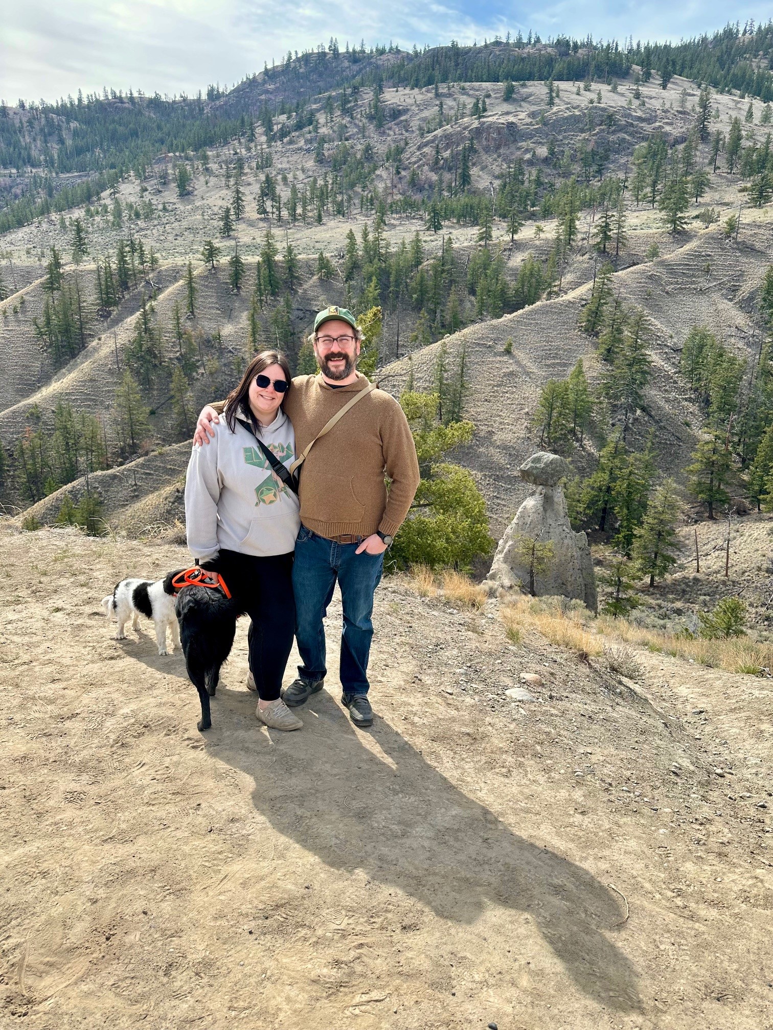 Woman and man standing with two dogs on a trail with forested hills in the background