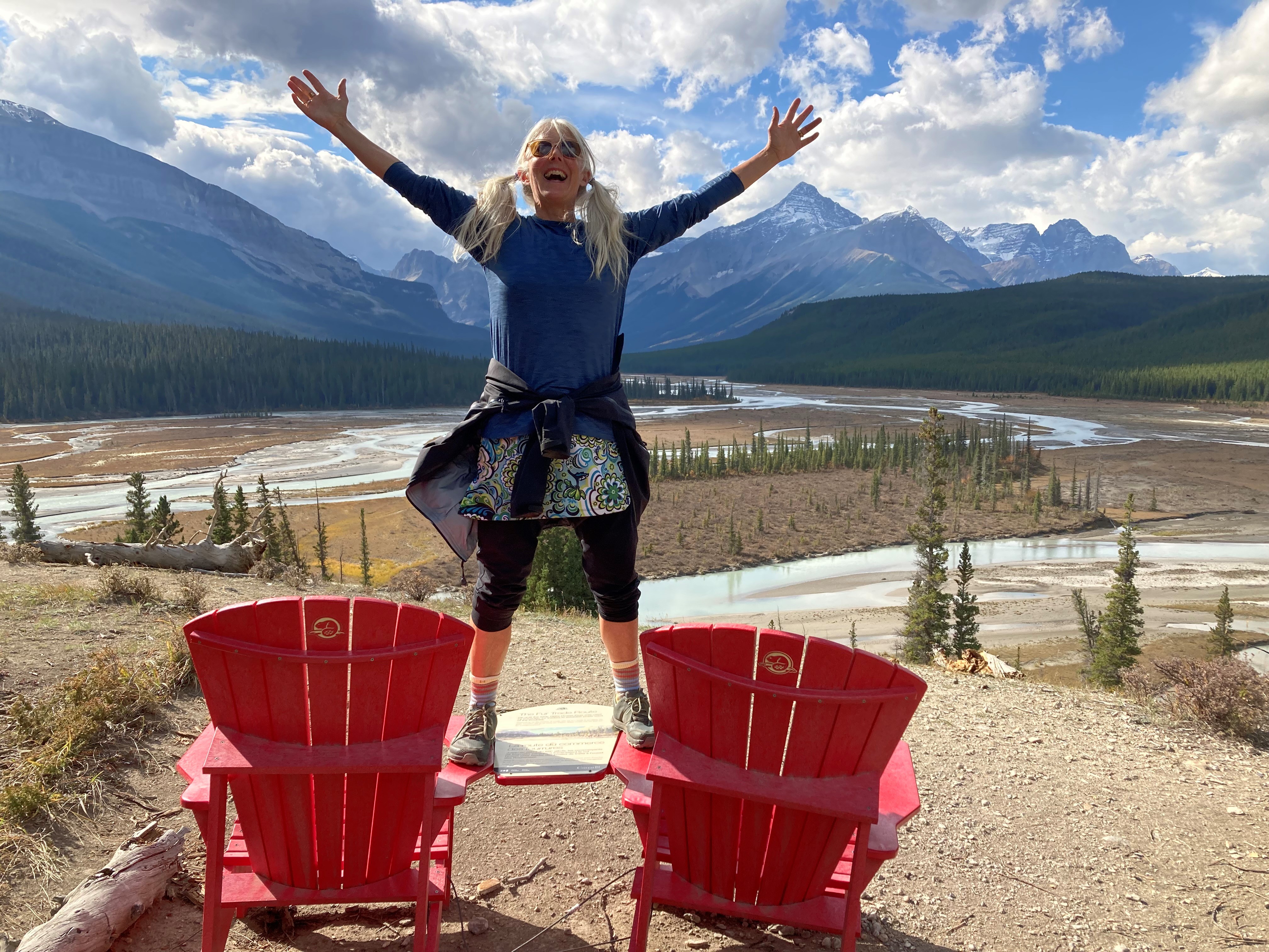 Woman with blonde pigtails standing on a table between two orange Adirondack chairs, arms raised, with mountains and a cloudy sky in the background.