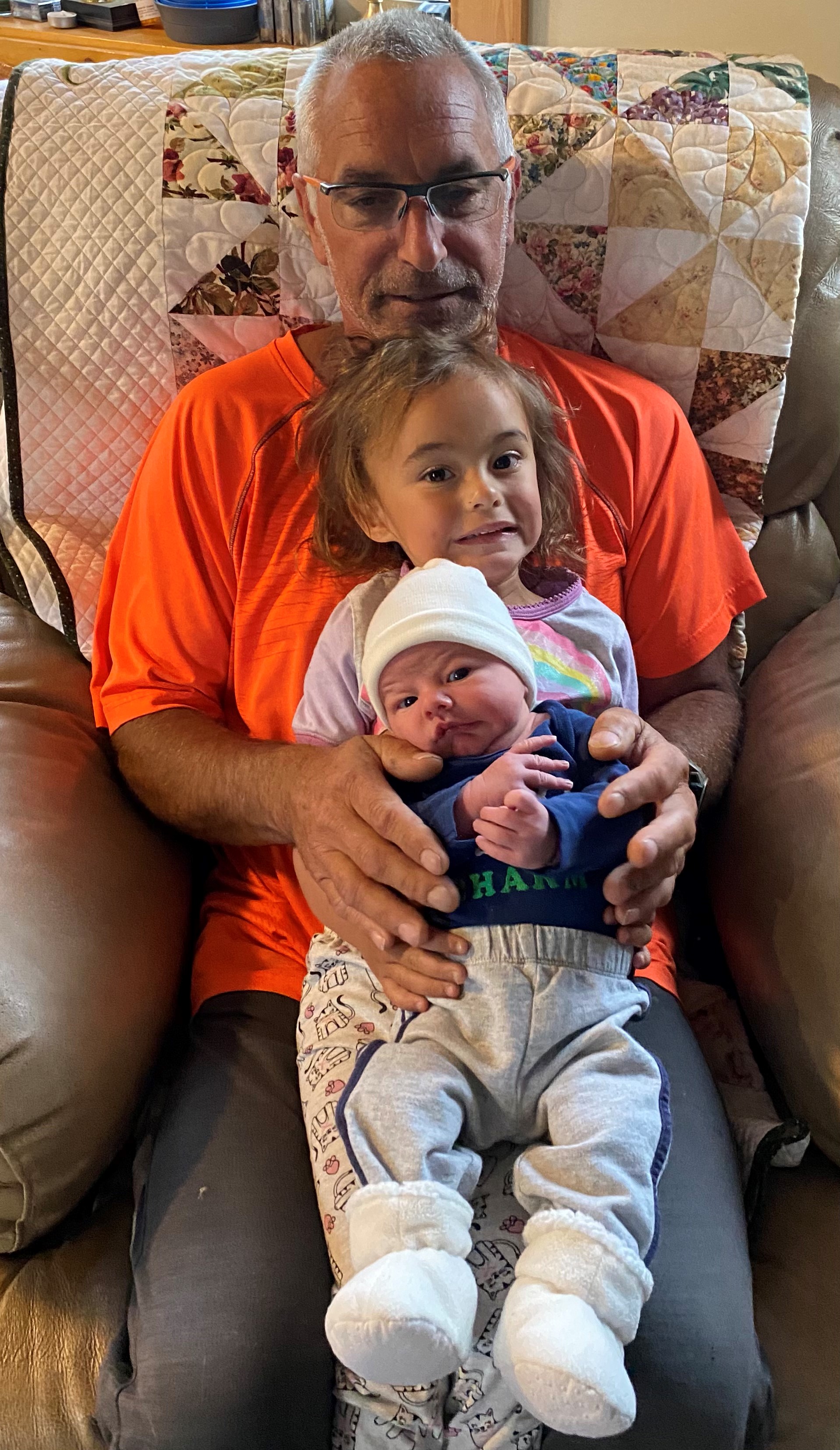 A man in an orange shift sitting on an armchair with a toddler and baby in his lap.