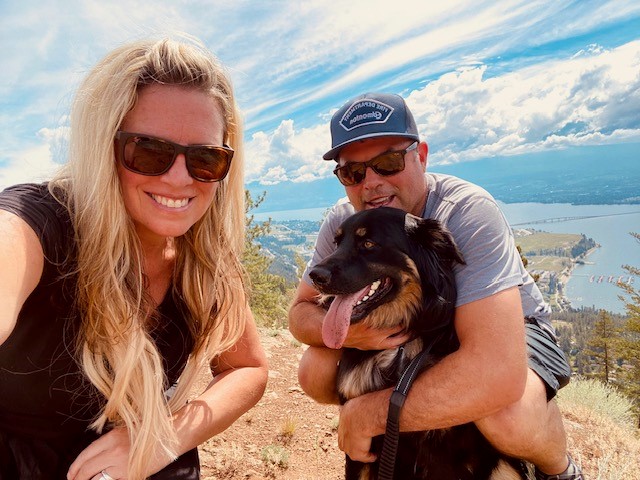 Smiling woman with long blonde hair wearing black top and sunglasses with man wearing blue ballcap, sunglasses and t-shirt, hugging dog, with lake, mountains and clouds in background