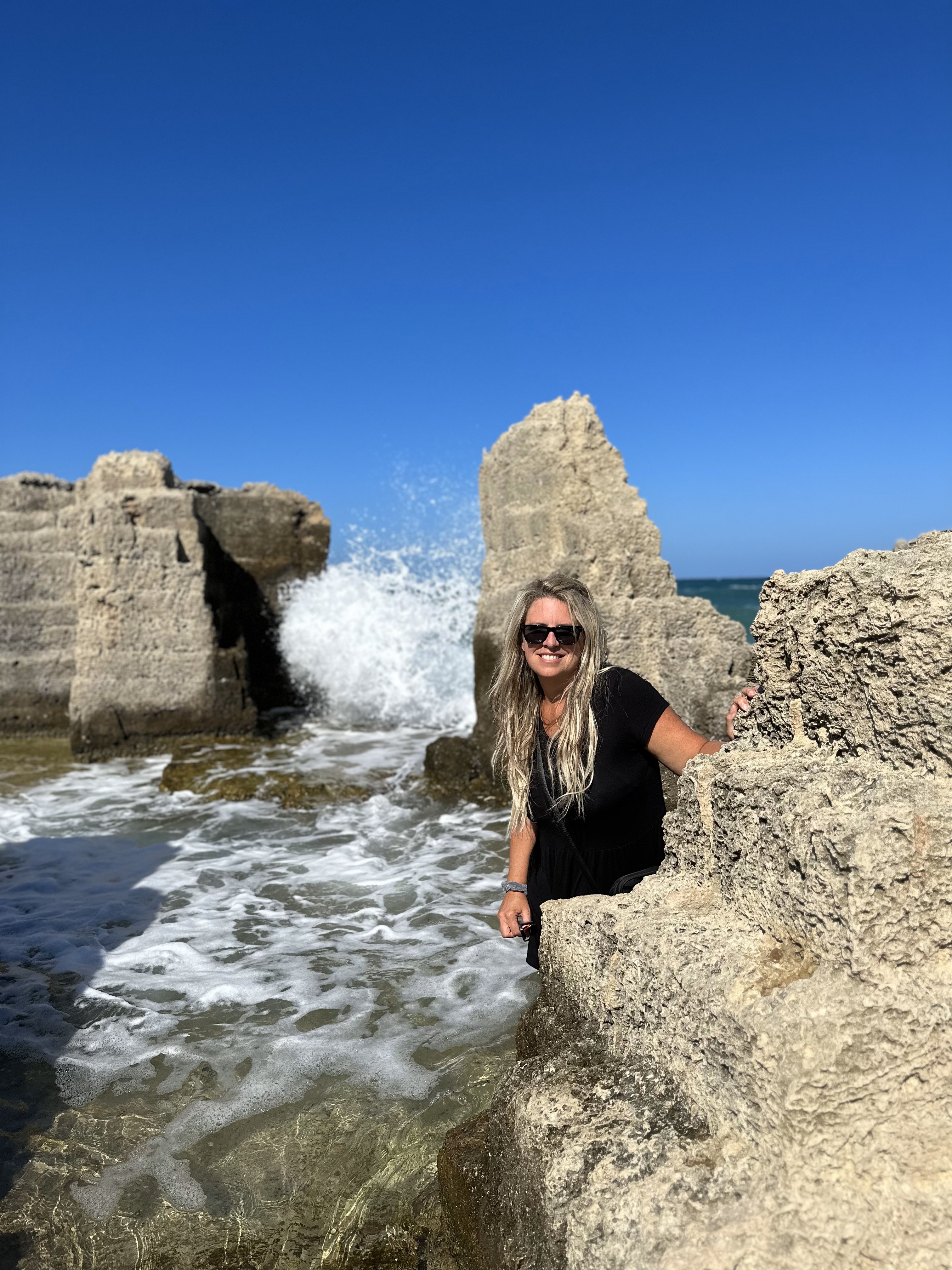 Smiling woman with long blonde hair wearing black top and sunglasses stands with stone wall