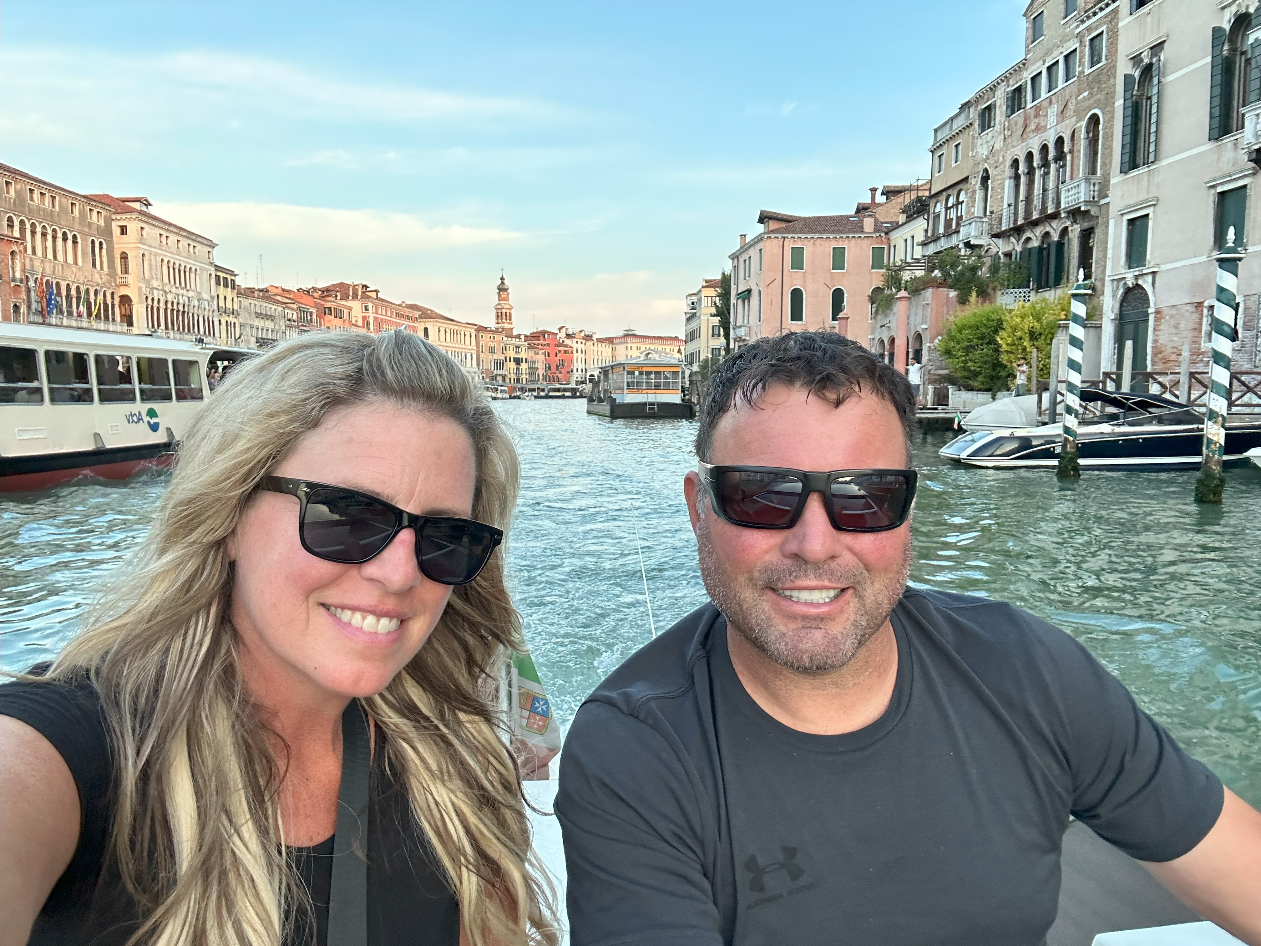 Smiling woman with long blonde hair wearing black top and sunglasses with man wearing grey t-shirt and sunglasses, with canal water, boats and houses in the background