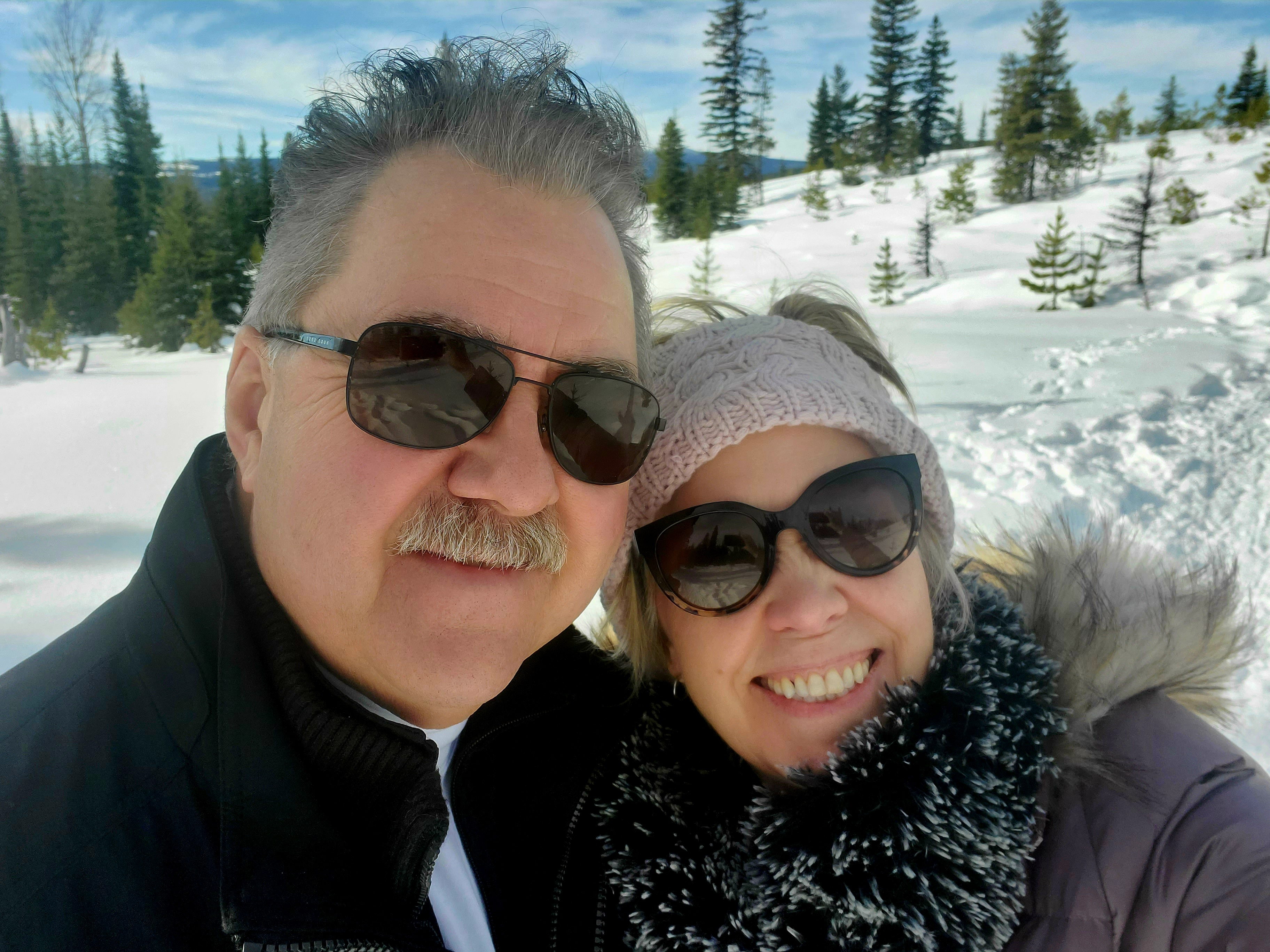 Two people: a woman with short blonde hair (right), wearing a winter coat and sunglasses stands next to a man with grey hair, wearing a winter jacket and sunglasses behind them is a snowy trail with green trees, and blue sky