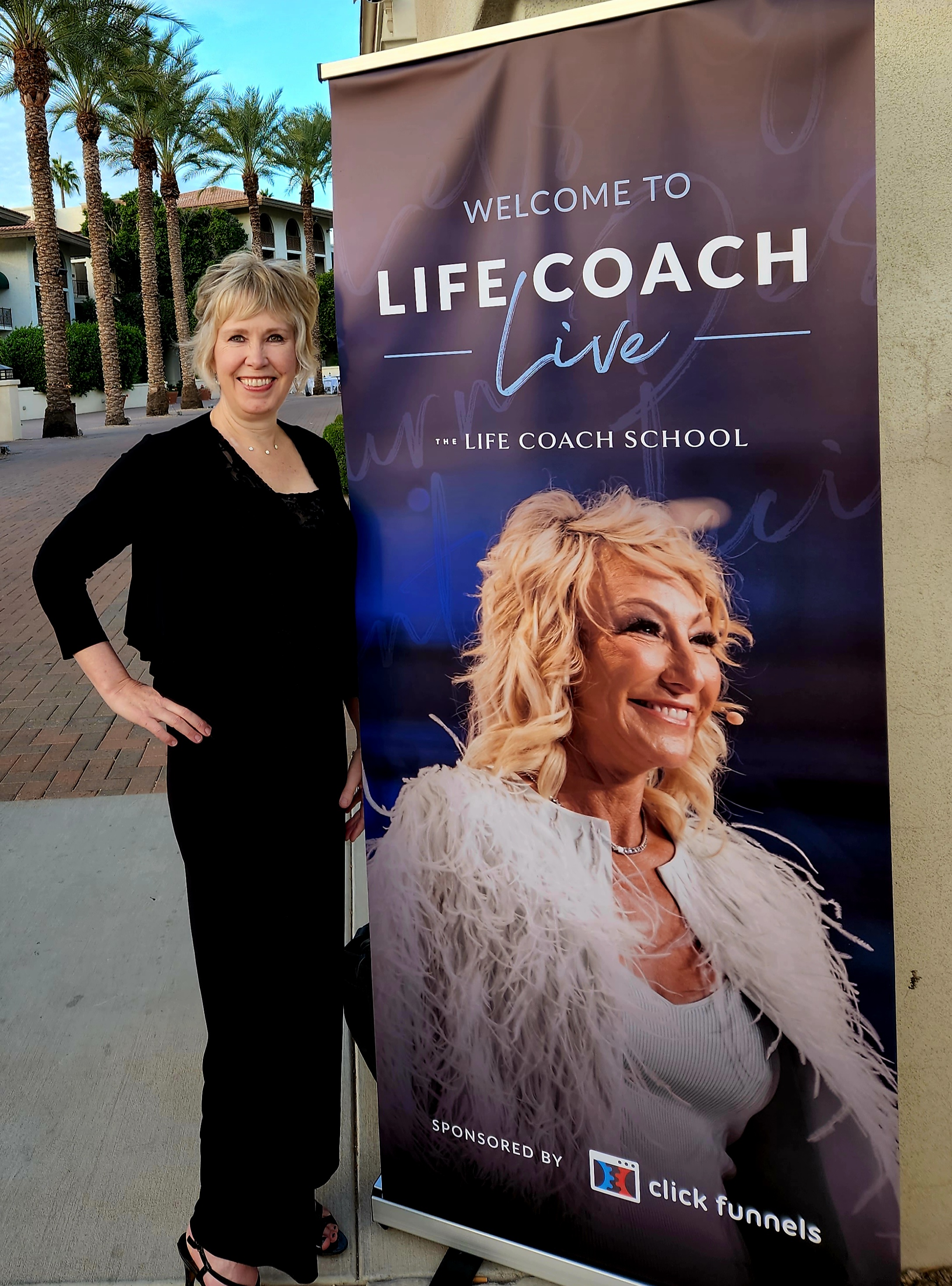 A woman with short blonde hair wearing all black stands next to large life coach event sign 