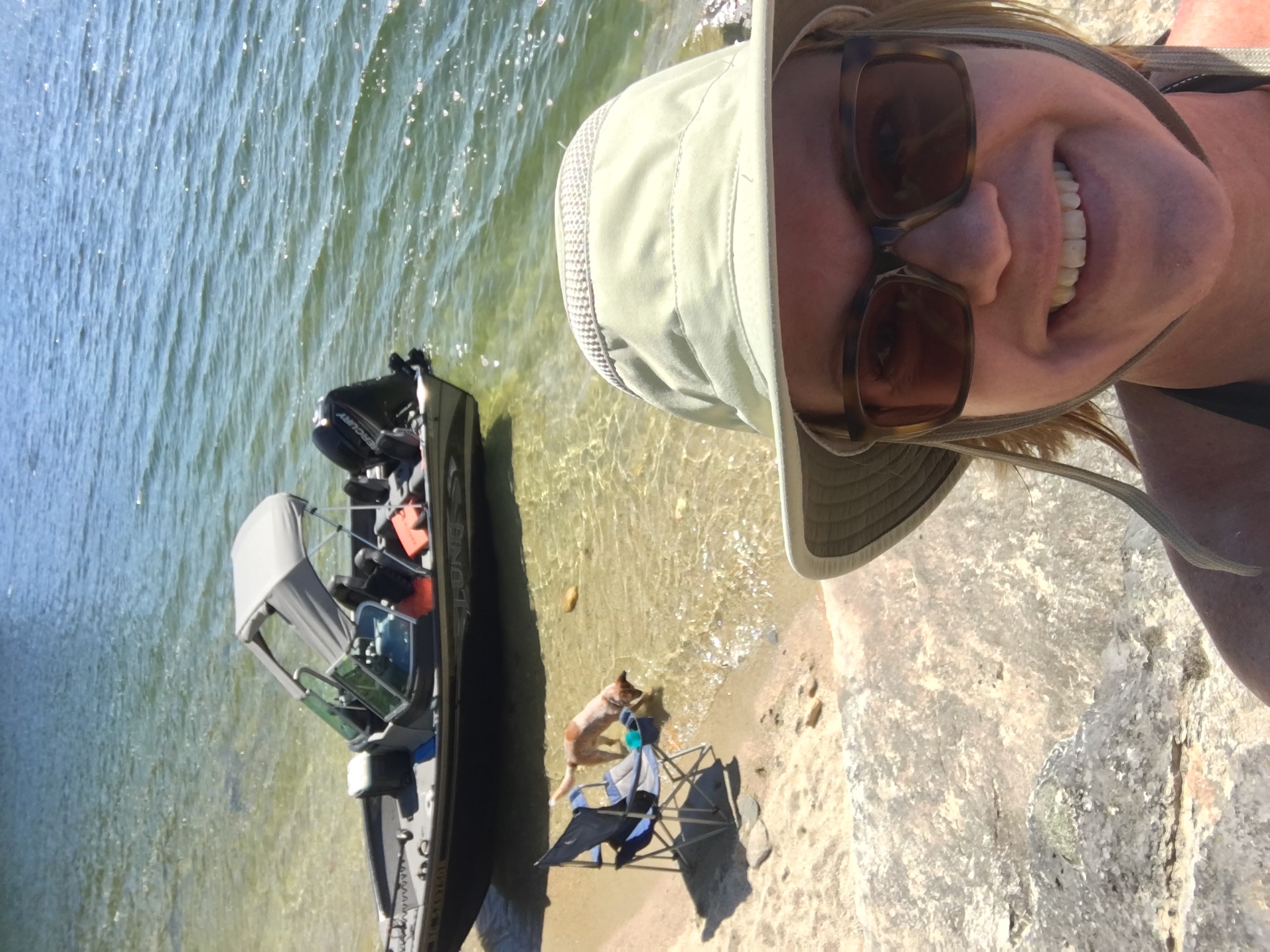 A woman takes a selfie near a lake with a dog and boat in the background