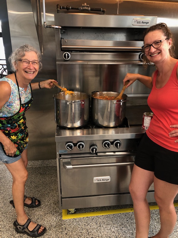 Two women, one older wearing a blue tank top and black apron, and one younger wearing a red tank top, stir two large stainless-steel pots of soup in a kitchen.