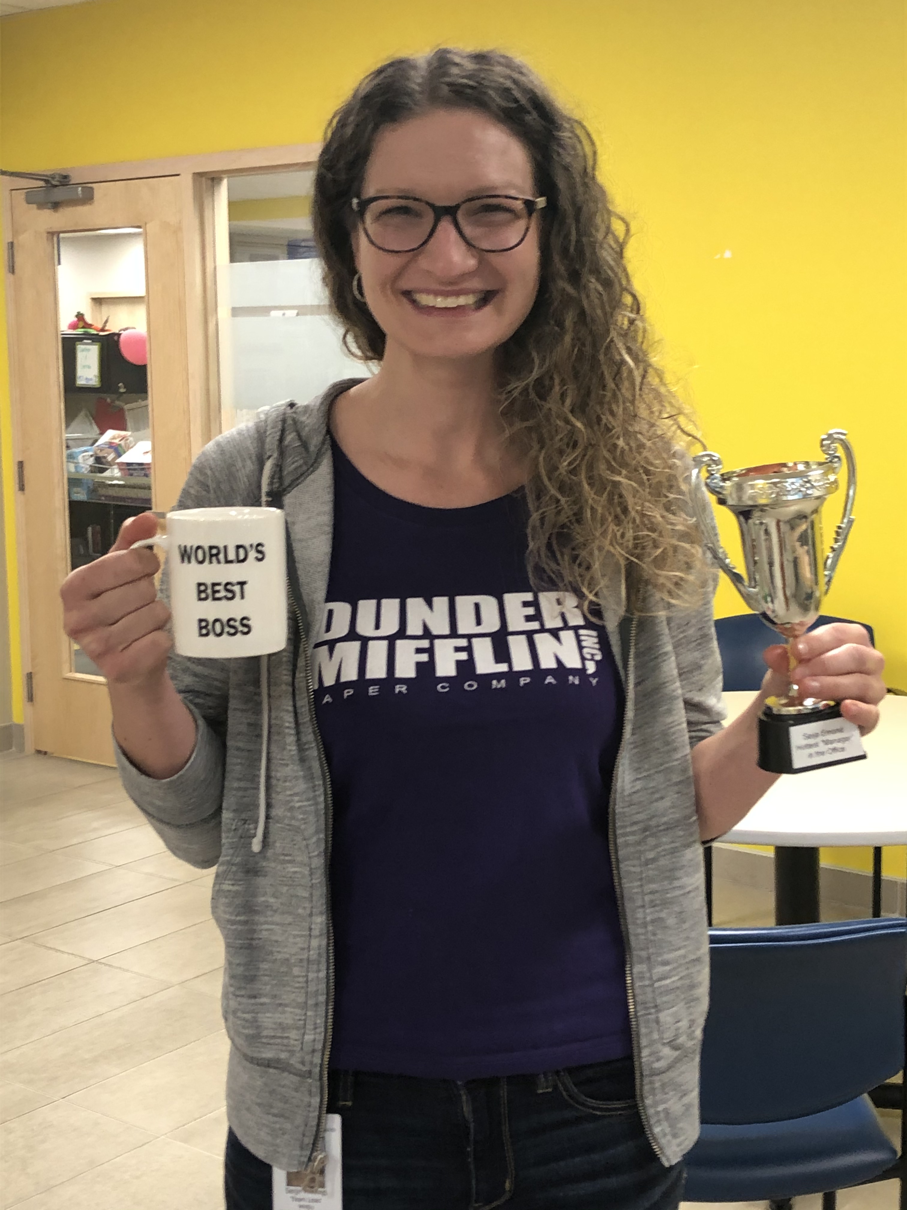 A smiling woman with long curly hair and glasses wearing a black T-shirt that says Dunder Mifflin holds a mug that says World’s Best Boss and a trophy in an office with yellow walls.