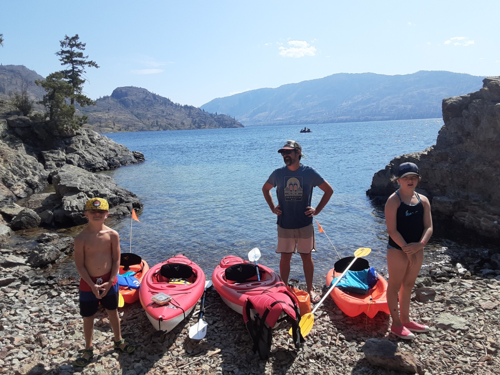 A man in a blue T-shirt and ballcap stands with his hands on his hips next to two young children in bathing suits next to two red kayaks and two orange kayaks on a rocky beach on a lake.