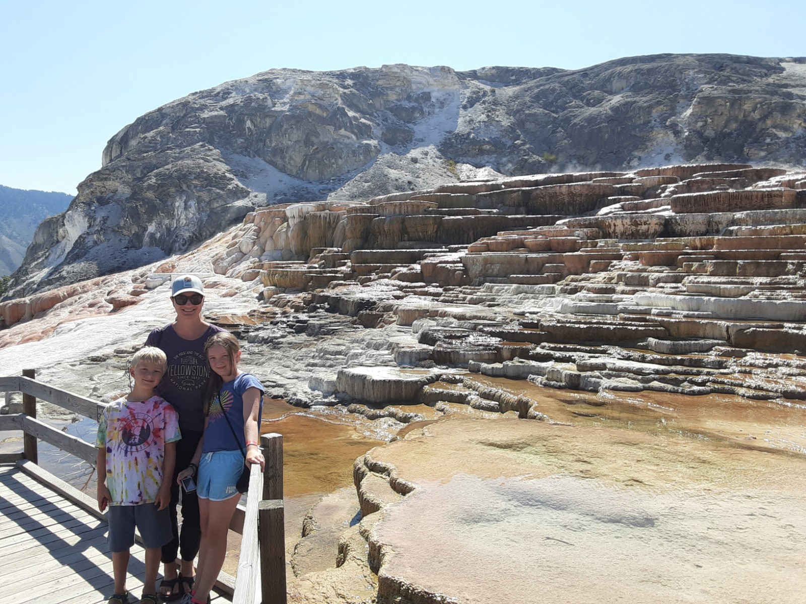 A person with a white ballcap and black T-shirt hugs two young children at a sunny location in front of a rocky outcrop and mountains