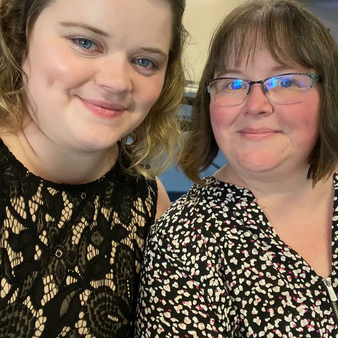 Mother wearing glasses and daughter, both with short hair and smiles, in black dresses with light-coloured pattern.