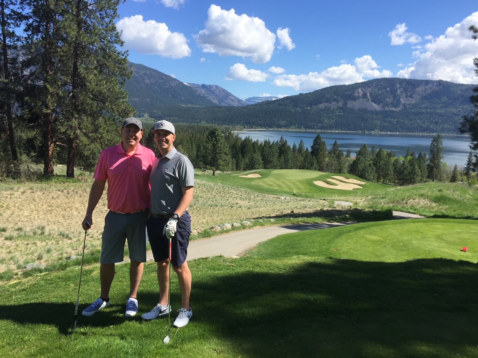 Two men standing on a golf green, with the golf course, trees and a lake behind them.