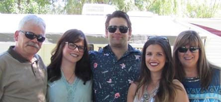 A family with their arms around each other. L-R: a man with grey hair, glasses and a moustache, a young woman with glasses and shoulder-length brown hair, a young man with short brown hair and black sunglasses, a young woman with long brown hair, an older woman with brown hair and dark sunglasses. 