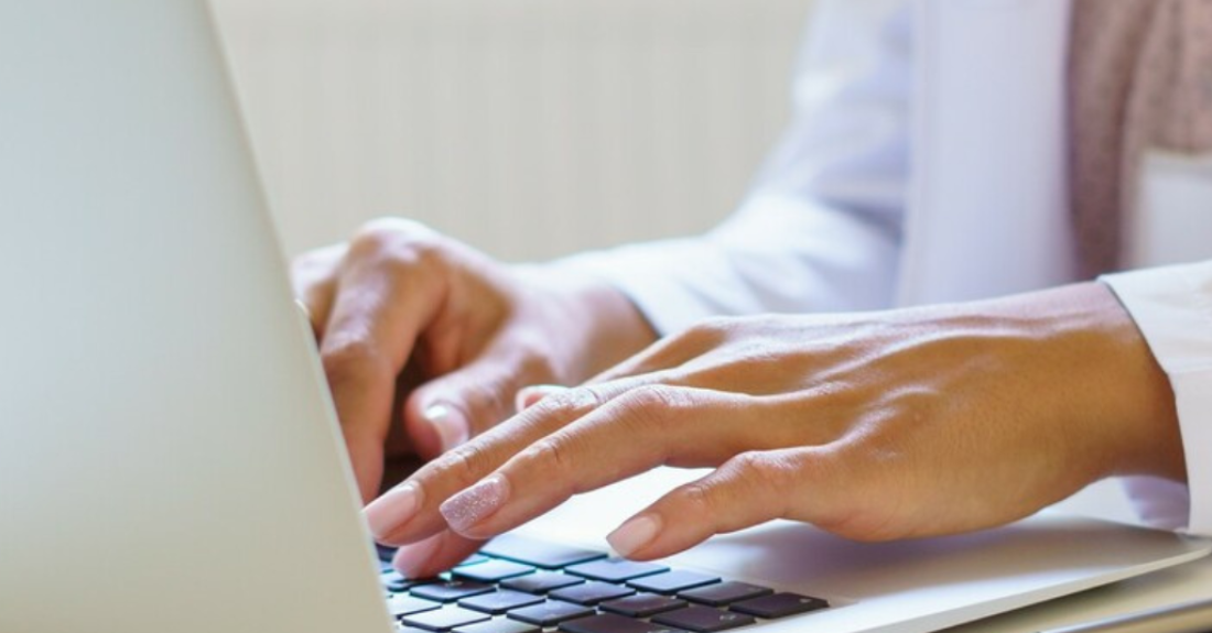 A pair of hands types on a laptop sitting on a beige table.