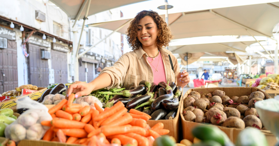 A smiling shopper at a market reaches for a carrot in a box surronded by other boxes with fresh vegetables.