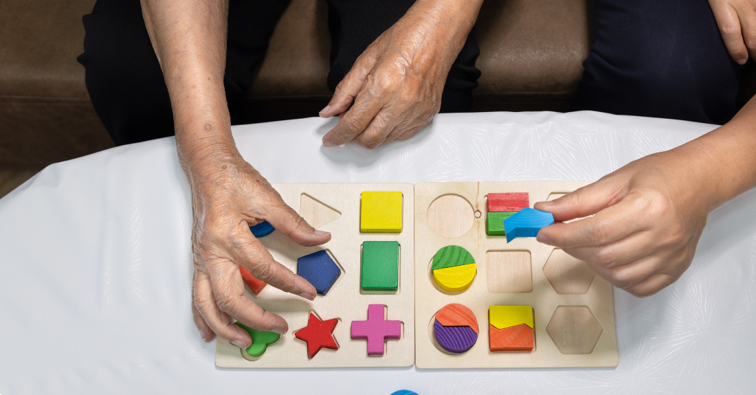 A pair of hands of an older person puts a coloured wooden shape into a space on a board