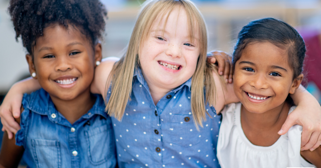 Three smiling girls arm in arm. The girl on the left has dark curly hair in a ponytail and bluejean shirt. The girl in the middle has long blonde hair and a blue polkdot shirt. The girl on the right has straight dark hair pulled back and is wearing a white blouse.