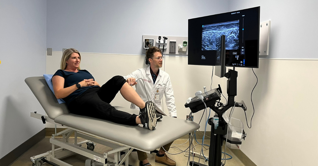 A patient lays on a medical bed as a health-care provider sitting to the side of the bed uses a small medical tool to scan their leg. They both look at a screen where an x-ray image is broadcasting the scan.