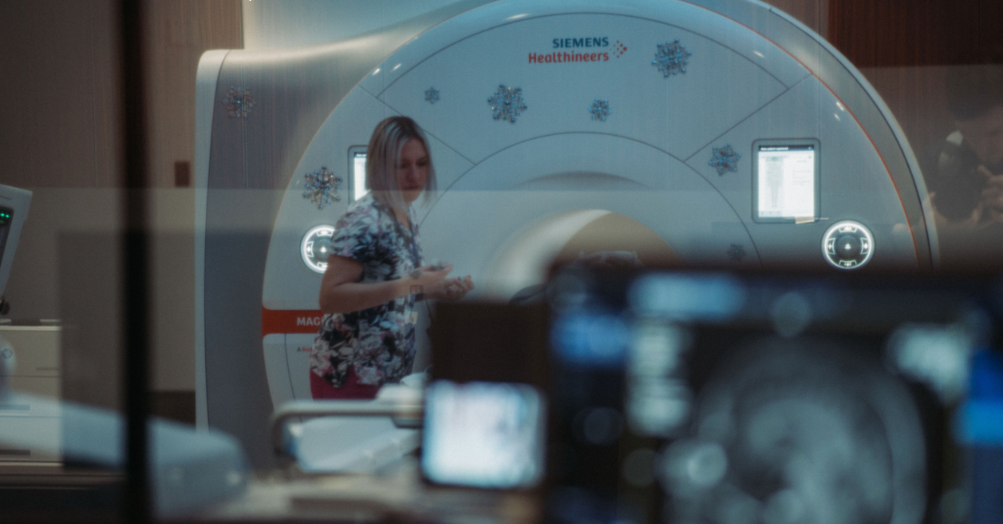 A person in medical clothing tends to a patient out of view in front of a large MRI machine with a blurred monitor in the foreground.